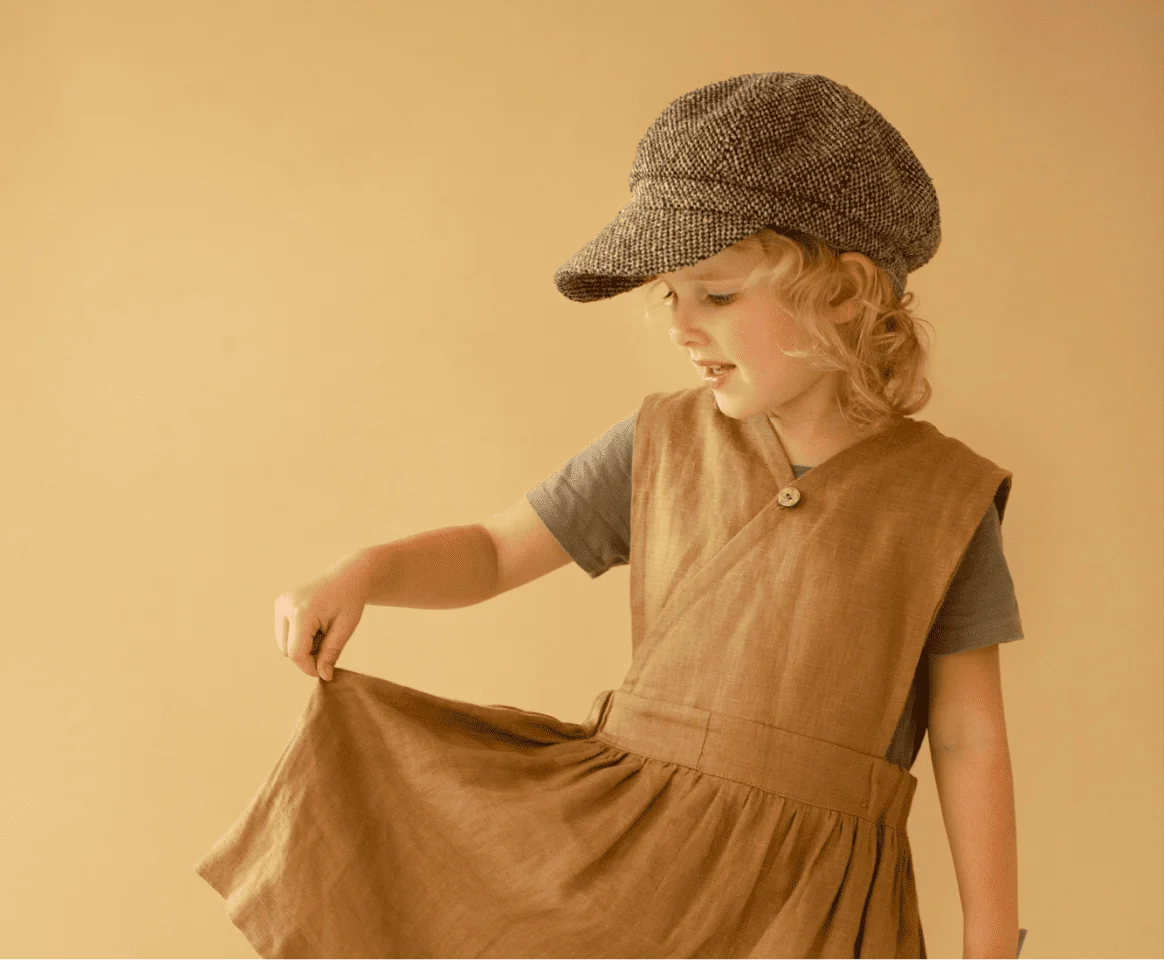 Toddler boy in a brown ribbed top standing by a curtain